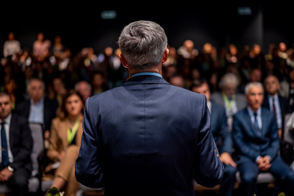 Business man is making a speech in front of a big audience at a conference hall. Speaker giving a talk on corporate business or political conference. Politician talking to group of people